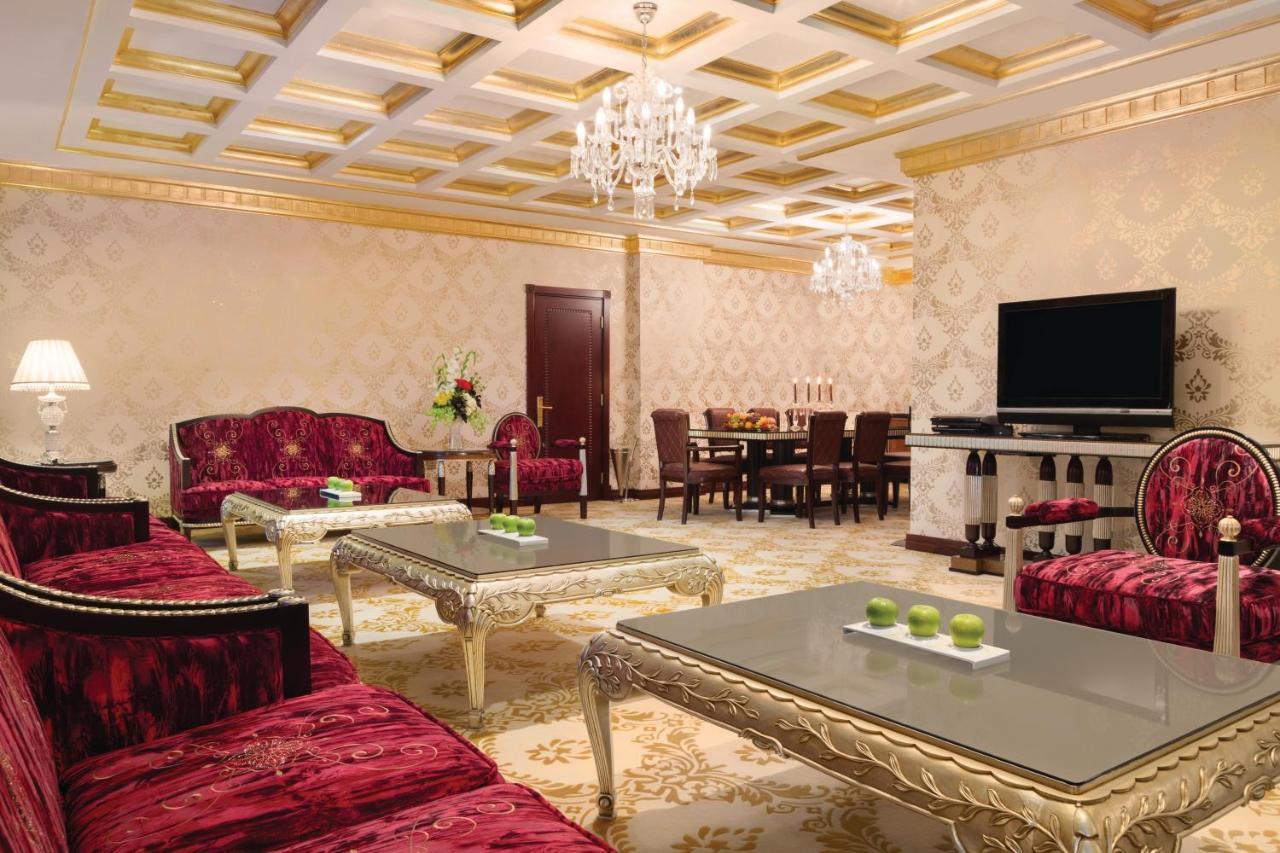 فندق ويندهام جراند ريجنسي الدوحة المظهر الخارجي الصورة The photo depicts an elegantly designed living room. The room features luxurious red sofas with ornate designs and metallic gold accents, arranged around glass-topped coffee tables that have intricate bases. A large, modern flat-screen television is 