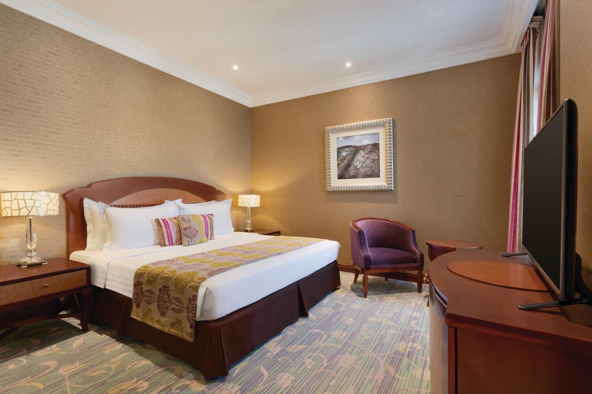 فندق ويندهام جراند ريجنسي الدوحة المظهر الخارجي الصورة The image shows a hotel room that features a cozy and elegant design. There's a large bed with white sheets and colorful decorative pillows. Next to the bed, there are nightstands with lamps. A comfortable armchair is positioned in the corner, and a 