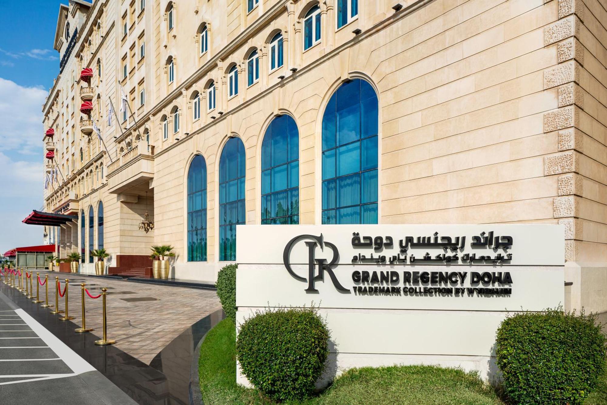 فندق ويندهام جراند ريجنسي الدوحة المظهر الخارجي الصورة The photo shows the entrance of the Grand Regency Doha, a luxury hotel. The building features a distinctive architectural style with large arched windows. There is a prominent sign featuring the hotel's name in both English and Arabic. The surroundin